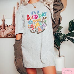 May include: A light gray t-shirt with the text "It's a good day to make some art" in colorful letters. The design includes a painter's palette with paintbrushes. The shirt is a casual, oversized style.