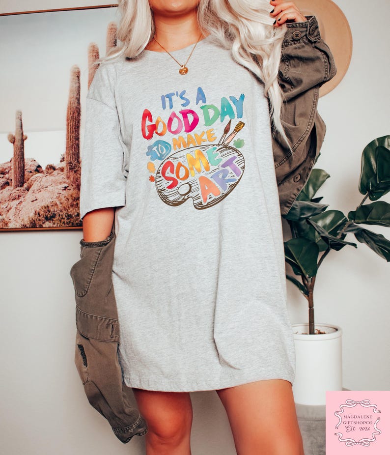 May include: A light gray t-shirt with the text "It's a good day to make some art" in colorful letters. The design includes a painter's palette with paintbrushes. The shirt is a casual, oversized style.