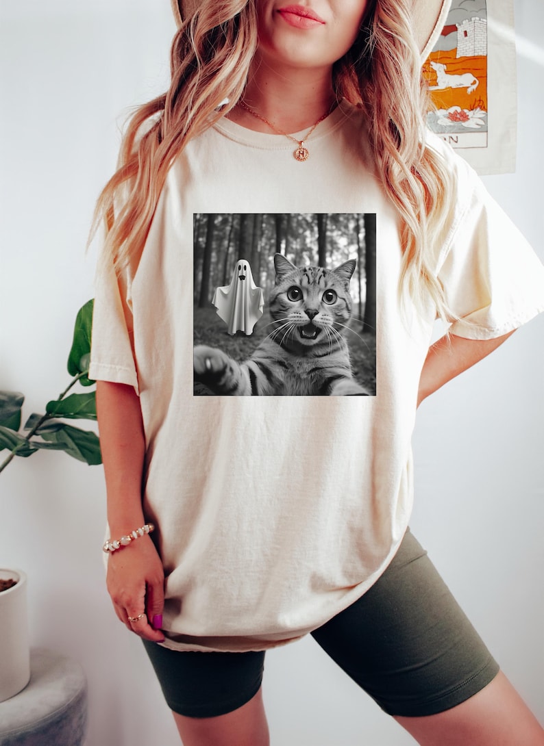 May include: Cream-colored t-shirt featuring a black and white image of a cat taking a selfie with a ghost in a forest. The cat has wide eyes and an open mouth. The shirt is a casual style.