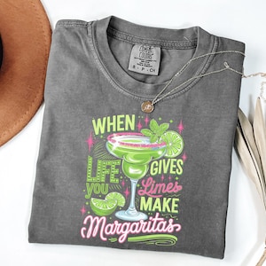 Könnte beinhalten: Graues T-Shirt mit einem Margarita-Motiv. Der Aufdruck lautet "When life gives you limes make margaritas" in Neon-Grün, Pink und Weiß. Eine silberne Halskette mit einem kleinen Anhänger befindet sich auf dem Shirt.