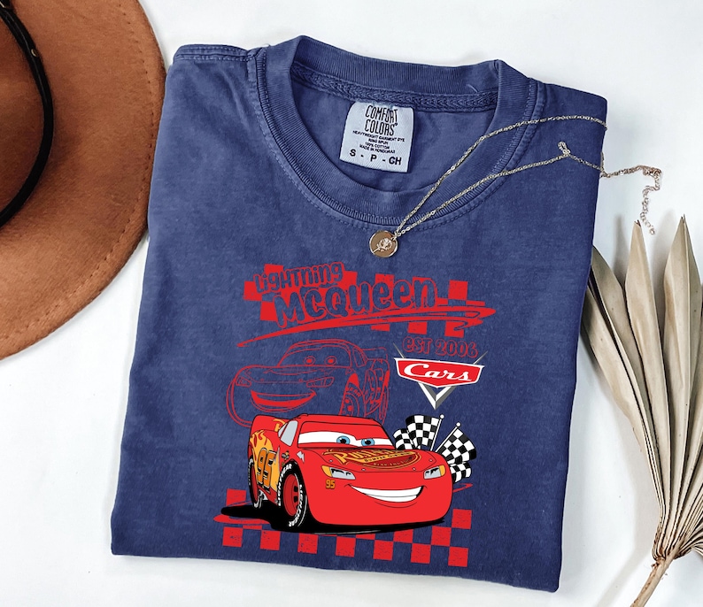 May include: A navy blue t-shirt with a graphic of Lightning McQueen from the Cars film. The design includes the text "Lightning McQueen EST 2006 Cars" in red, and checkered flags. A gold necklace with a pendant is draped across the shirt.