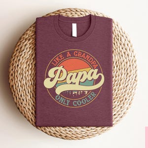 May include: A maroon t-shirt with a circular graphic that reads "Like a Grandpa Papa Only Cooler" in a retro font. The graphic features a sunset design with orange, yellow, and teal colors. The shirt is folded on a woven surface.