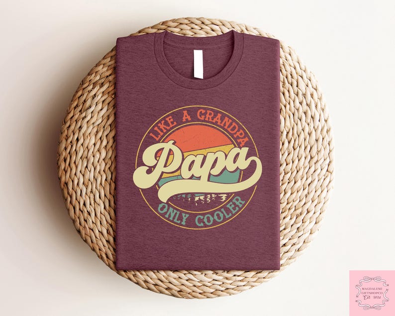 May include: A maroon t-shirt with a circular graphic that reads "Like a Grandpa Papa Only Cooler" in a retro font. The graphic features a sunset design with orange, yellow, and teal colors. The shirt is folded on a woven surface.