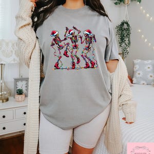 May include: A gray t-shirt features a graphic of three dancing skeletons wearing Santa hats and red boots, strung with colorful Christmas lights. The shirt is part of a holiday-themed collection.
