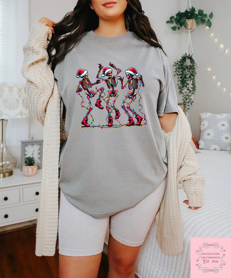 May include: A gray t-shirt features a graphic of three dancing skeletons wearing Santa hats and red boots, strung with colorful Christmas lights. The shirt is part of a holiday-themed collection.