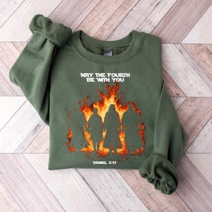 K&ouml;nnte beinhalten: Olivgr&uuml;nes Sweatshirt mit dem Text "MAY THE FOURTH BE WITH YOU" &uuml;ber einer Grafik von Silhouetten vor Flammen. Der Text "DANIEL 3:17" steht darunter. Das Sweatshirt liegt gefaltet auf einer hellen Holzoberfl&auml;che.
