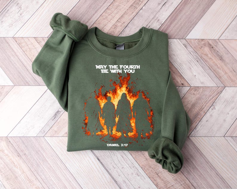 K&ouml;nnte beinhalten: Olivgr&uuml;nes Sweatshirt mit dem Text "MAY THE FOURTH BE WITH YOU" &uuml;ber einer Grafik von Silhouetten vor Flammen. Der Text "DANIEL 3:17" steht darunter. Das Sweatshirt liegt gefaltet auf einer hellen Holzoberfl&auml;che.