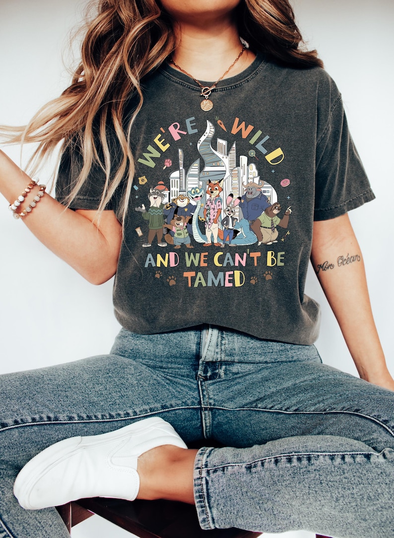 May include: Dark gray t-shirt with the text "WE'RE WILD AND WE CAN'T BE TAMED" above a colorful cartoon illustration of animal characters in a city setting. The shirt is worn with blue jeans and white sneakers.