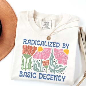 以下が含まれることがあります： 「RADICALIZED BY BASIC DECENCY」の文字が青色で、ピンク、オレンジ、黄色の花柄がプリントされたクリーム色のTシャツ。 金色のネックレスがシャツの上に置かれています。 シャツはComfort Colorsブランドです。