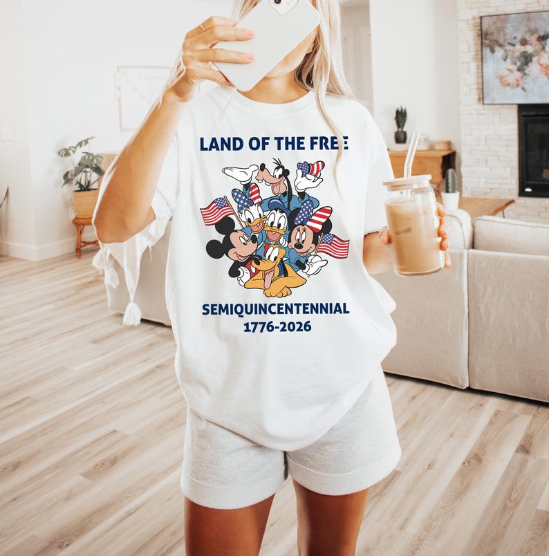 May include: A white t-shirt featuring a graphic of cartoon characters including Mickey Mouse, Minnie Mouse, Donald Duck, Goofy, and Pluto wearing patriotic hats and holding American flags. The text reads LAND OF THE FREE SEMIQUINCENTENNIAL 1776-2026.