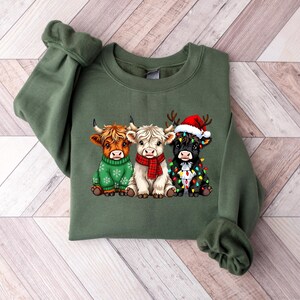 Pu&ograve; includere: Una felpa verde oliva con una grafica di tre mucche delle Highlands. Le mucche sono adornate con abiti festivi: un maglione verde, una sciarpa rossa e un cappello di Babbo Natale con luci natalizie. La felpa ha maniche lunghe.