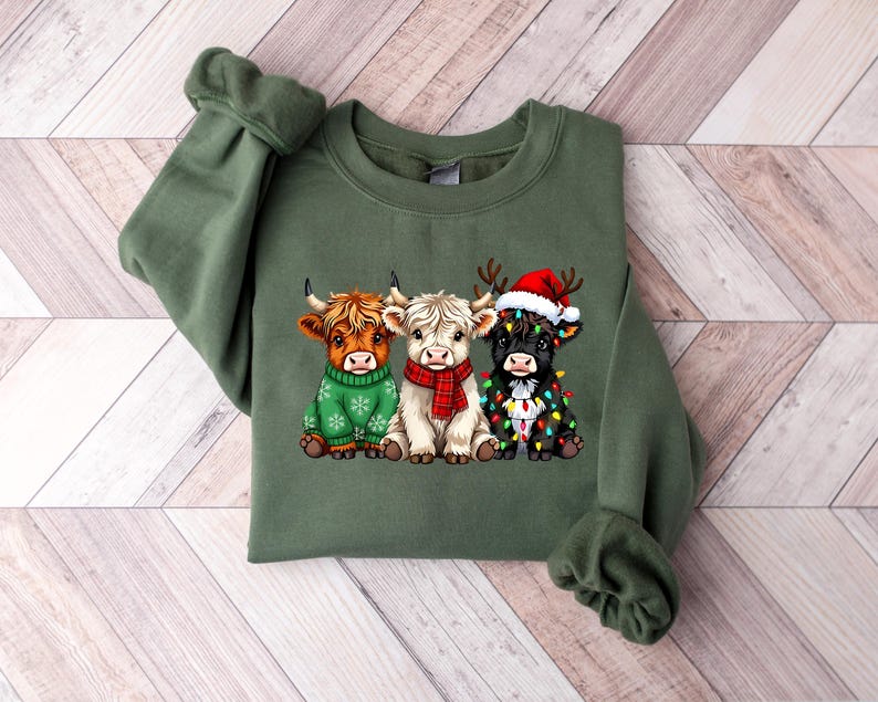 Pu&ograve; includere: Una felpa verde oliva con una grafica di tre mucche delle Highlands. Le mucche sono adornate con abiti festivi: un maglione verde, una sciarpa rossa e un cappello di Babbo Natale con luci natalizie. La felpa ha maniche lunghe.