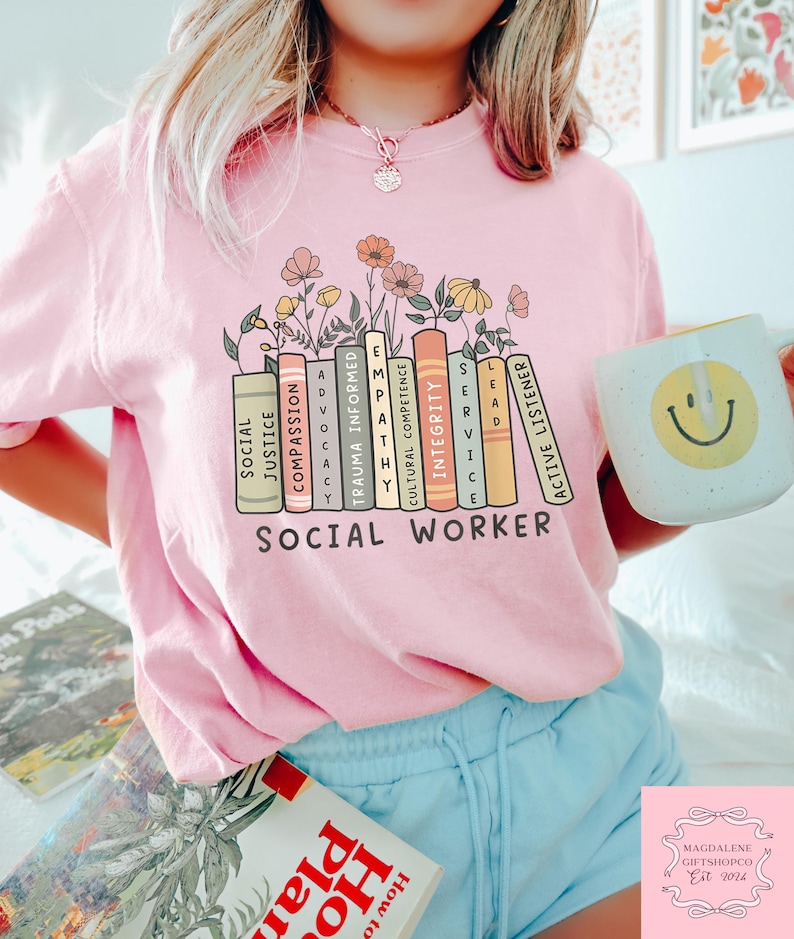 Pu&ograve; includere: Maglietta rosa con una grafica di libri impilati con parole come "Giustizia sociale", "Compassione", "Empatia", "Informato sul trauma", "Competenza culturale", "Integrit&agrave;", "Leadership", "Servizio" e "Ascolto attivo". Il testo "Assistente sociale" &egrave; stampato sotto la grafica.