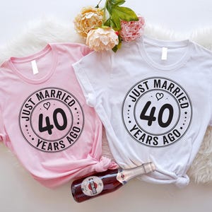 Op de afbeelding: Twee roze en witte T-shirts met een zwart cirkelontwerp met de tekst "Just Married 40 Years Ago".