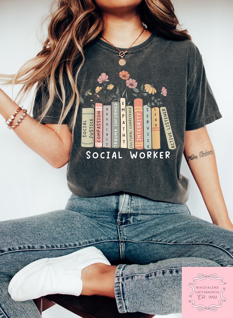 Pu&ograve; includere: Una maglietta grigio scuro con una grafica di libri impilati uno sopra l'altro. I libri hanno parole sui dorsi, tra cui "Giustizia sociale", "Compassione", "Empatia", "Informato sul trauma", "Competenza culturale", "Integrit&agrave;", "Guida", "Servizio", e "Ascolto attivo". Il testo "LAVORATORE SOCIALE" &egrave; stampato sotto i libri.
