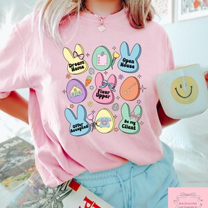 May include: A pink t-shirt with a graphic design of pastel colored Easter eggs with bunny ears and text that says "Dream Home", "Open House", "Fixer Upper", "Offer Accepted", "Be My Client", and "Magdalene Giftshopco Est 2014".