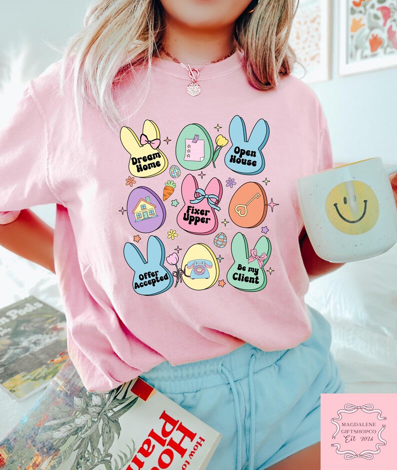 May include: A pink t-shirt with a graphic design of pastel colored Easter eggs with bunny ears and text that says "Dream Home", "Open House", "Fixer Upper", "Offer Accepted", "Be My Client", and "Magdalene Giftshopco Est 2014".