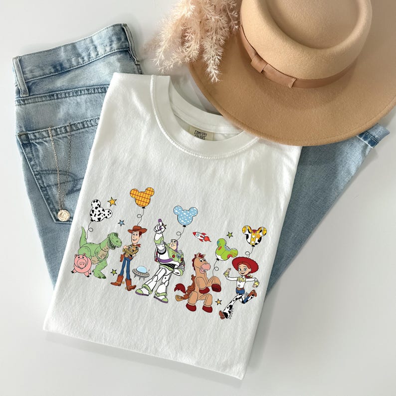 May include: White crew neck t-shirt featuring a colorful graphic of animated characters with balloons. The shirt is paired with light wash denim jeans and a tan fedora hat. A gold necklace is visible.