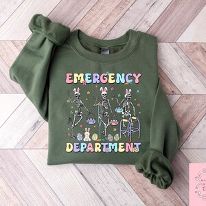 May include: Green sweatshirt with a graphic of three skeletons holding Easter eggs and walking aids. The text "Emergency Department" is written in colorful letters.
