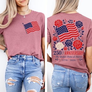 以下が含まれることがあります： 前面と背面にアメリカ国旗がプリントされたモーヴ色のTシャツ。前面には小さな旗のグラフィックがあり、背面には赤、白、青の花柄と「250th Anniversary The United States of America 1776-2026」の文字で囲まれた大きな旗のデザインが表示されています。