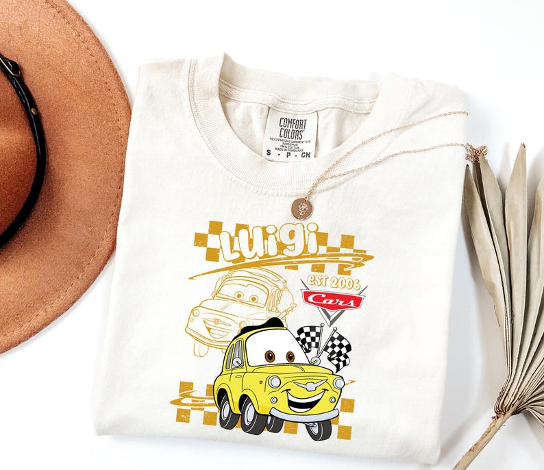 May include: Cream-coloured t-shirt featuring a yellow cartoon car named Luigi from the movie Cars. The shirt has the text "Luigi" in gold, along with the Cars logo and checkered flags. A gold necklace is draped over the shirt.