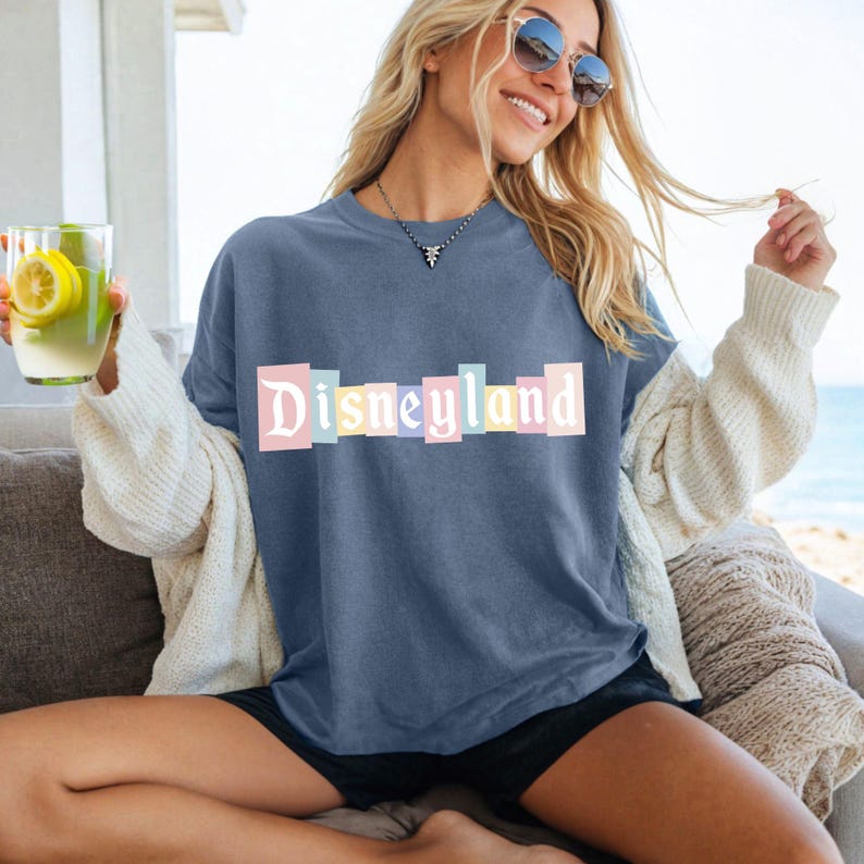 May include: A blue t-shirt with the word "Disneyland" in pastel-colored block letters. The shirt is worn with black shorts and a cream-colored cardigan. The person is sitting on a couch near the beach.