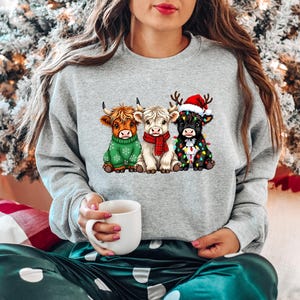 Pu&ograve; includere: Felpa grigio chiaro con una grafica di tre mucche delle Highlands vestite per Natale. Una mucca indossa un maglione verde, un'altra una sciarpa rossa e la terza un cappello da Babbo Natale e luci natalizie. La persona tiene in mano una tazza bianca.