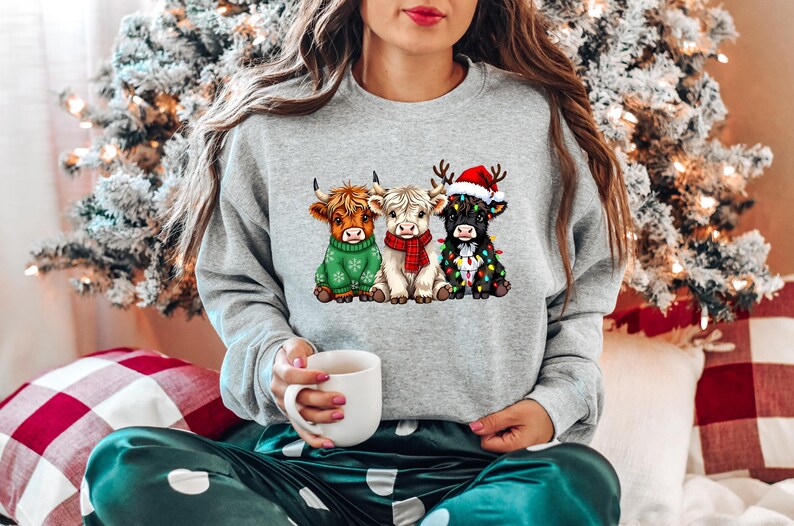 Pu&ograve; includere: Felpa grigio chiaro con una grafica di tre mucche delle Highlands vestite per Natale. Una mucca indossa un maglione verde, un'altra una sciarpa rossa e la terza un cappello da Babbo Natale e luci natalizie. La persona tiene in mano una tazza bianca.