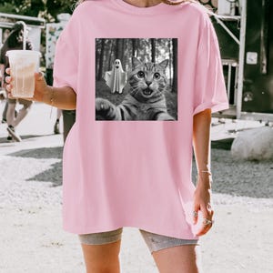 May include: A light pink oversized t-shirt featuring a black and white graphic of a cat taking a selfie with a ghost in a forest. The cat has wide eyes and an open mouth. The shirt has short sleeves.