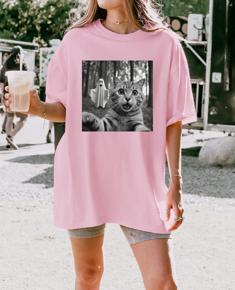 May include: A light pink oversized t-shirt featuring a black and white graphic of a cat taking a selfie with a ghost in a forest. The cat has wide eyes and an open mouth. The shirt has short sleeves.