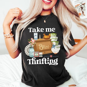 May include: Black t-shirt with a graphic design featuring a bread box, bowls, a duck, and the text "Take me Thrifting." The design includes a mason jar, flowers, and other vintage-style elements. The shirt is a comfortable, casual style.