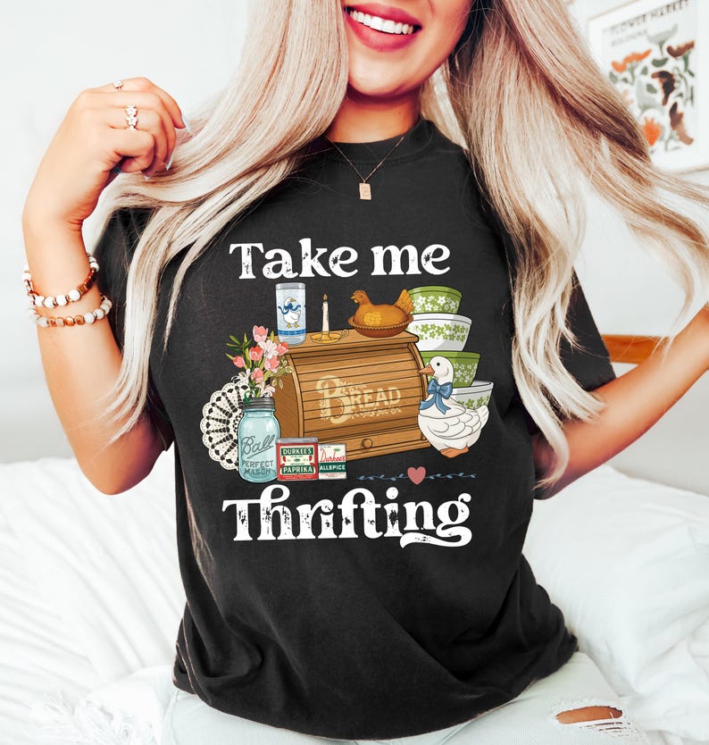 May include: Black t-shirt with a graphic design featuring a bread box, bowls, a duck, and the text "Take me Thrifting." The design includes a mason jar, flowers, and other vintage-style elements. The shirt is a comfortable, casual style.