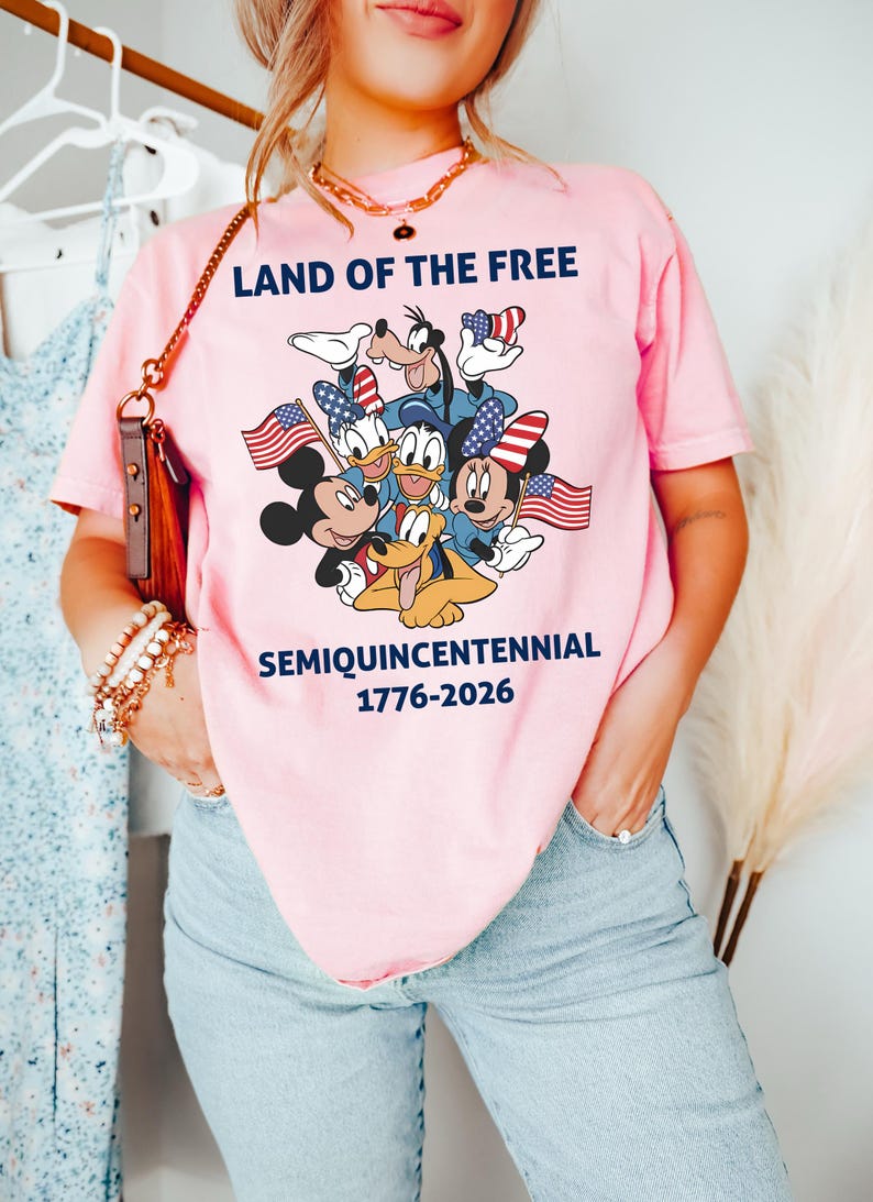 May include: A light pink short-sleeve t-shirt featuring a graphic of cartoon characters holding American flags. The text reads LAND OF THE FREE, SEMIQUINCENTENNIAL, and 1776-2026 in dark blue lettering. The shirt is paired with light blue denim jeans.