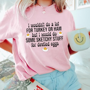 May include: A light pink sweatshirt with the text "I wouldn't do a lot for turkey or ham but I would do some sketchy stuff for deviled eggs." A white mug with a yellow smiley face is held in the hand.