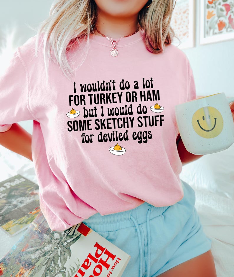 May include: A light pink sweatshirt with the text "I wouldn't do a lot for turkey or ham but I would do some sketchy stuff for deviled eggs." A white mug with a yellow smiley face is held in the hand.