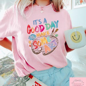 May include: A light pink t-shirt with the text "IT'S A GOOD DAY TO MAKE SOME ART" in colorful letters and a painter's palette design. A white mug with a yellow smiley face is held in the hand. The person is wearing blue shorts.