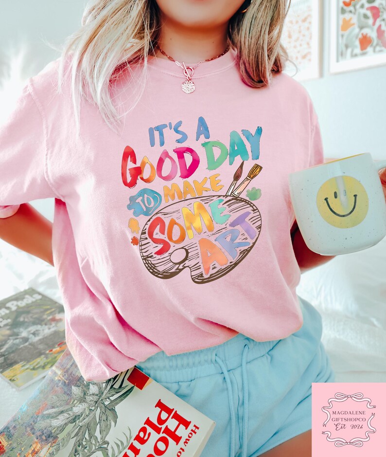 May include: A light pink t-shirt with the text "IT'S A GOOD DAY TO MAKE SOME ART" in colorful letters and a painter's palette design. A white mug with a yellow smiley face is held in the hand. The person is wearing blue shorts.
