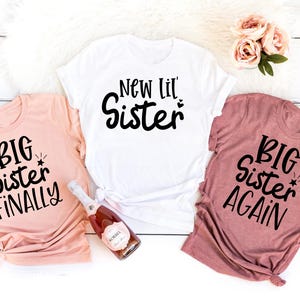 Puede incluir: Tres camisetas con texto impreso. La primera camiseta es rosa y dice "Big Sister Finally". La segunda camiseta es blanca y dice "New Lil' Sister". La tercera camiseta es de color marrón claro y dice "Big Sister Again".