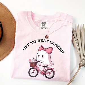 以下が含まれることがあります： ピンク色のTシャツ。ピンクのリボンをつけた白いお化けが、花かごを乗せたピンクの自転車に乗っているイラストが描かれています。イラストの上には黒い大文字で「OFF TO BEAT CANCER」と書かれています。Comfort Colorsのタグ付き。