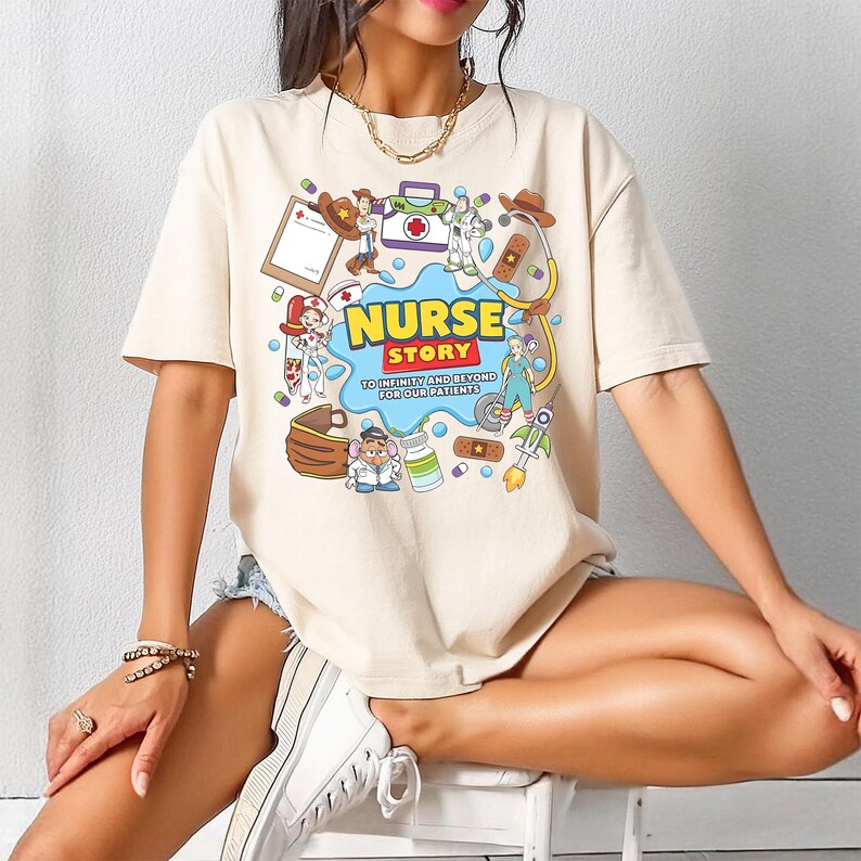 Pu&ograve; includere: T-shirt beige con una grafica a cartoni animati colorata. La grafica presenta le parole "NURSE STORY" in giallo e blu, circondate da illustrazioni a tema medico e il testo "TO INFINITY AND BEYOND FOR OUR PATIENTS."
