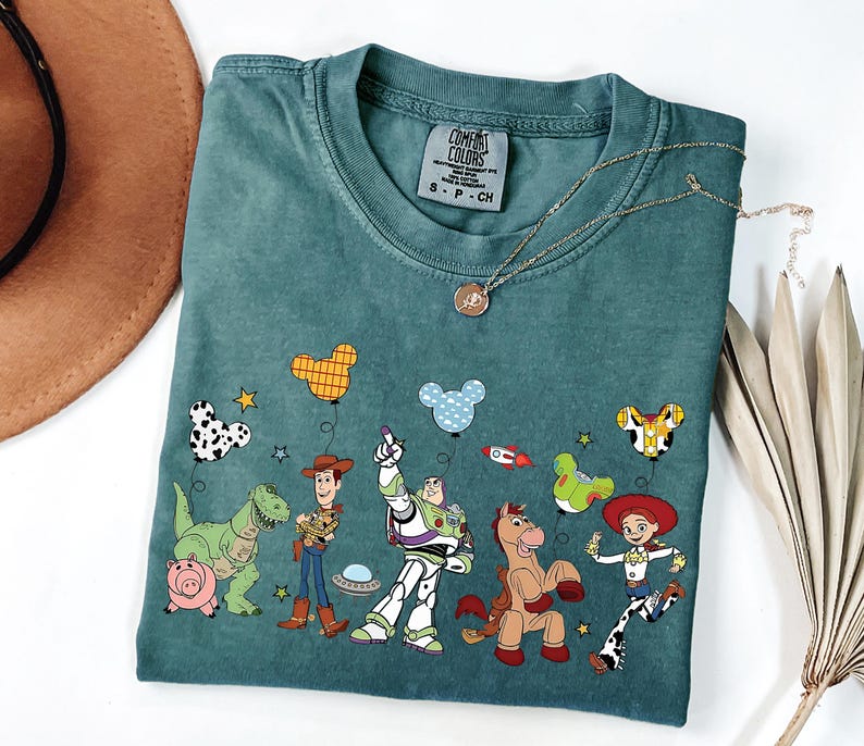 May include: Teal Comfort Colors t-shirt featuring a colorful graphic of Toy Story characters and balloons. The design includes Woody, Buzz Lightyear, Jessie, and other characters. A gold necklace with a pendant is draped on the shirt.