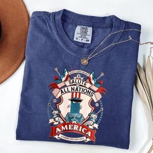 May include: A navy blue Comfort Colors t-shirt featuring a graphic that reads "A Salute to All Nations, Mostly America." The graphic includes a cartoon character in a top hat, flags, and banners. A gold necklace with a pendant is draped on the shirt.