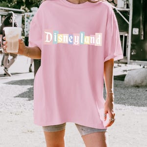 May include: A light pink t-shirt with the word "Disneyland" printed across the front in pastel colors. The shirt is a loose fit and appears to be made of a soft material. The overall style is casual and relaxed.