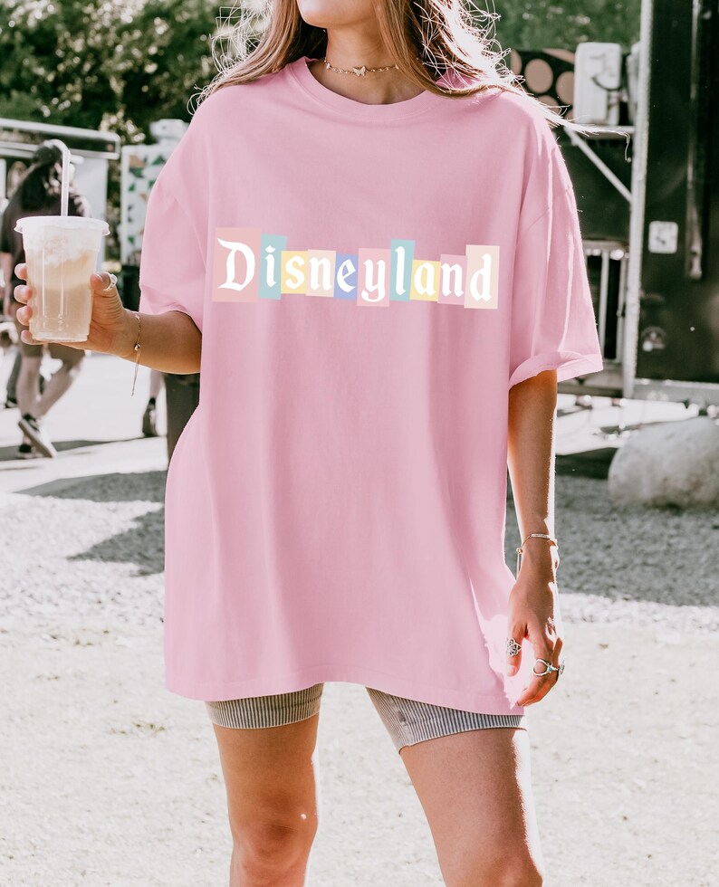 May include: A light pink t-shirt with the word "Disneyland" printed across the front in pastel colors. The shirt is a loose fit and appears to be made of a soft material. The overall style is casual and relaxed.