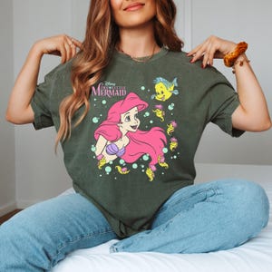 Puede incluir: Camiseta verde oscuro con un gr&aacute;fico de Ariel de La Sirenita de Disney. Ariel tiene el pelo rosa y un sujetador de concha p&uacute;rpura. La camiseta tambi&eacute;n presenta a Flounder y caballitos de mar. El texto "The Little Mermaid" est&aacute; encima de Ariel.