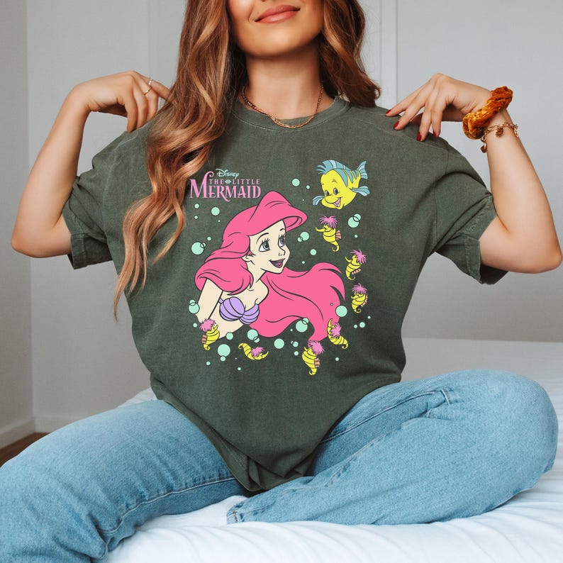 Puede incluir: Camiseta verde oscuro con un gr&aacute;fico de Ariel de La Sirenita de Disney. Ariel tiene el pelo rosa y un sujetador de concha p&uacute;rpura. La camiseta tambi&eacute;n presenta a Flounder y caballitos de mar. El texto "The Little Mermaid" est&aacute; encima de Ariel.