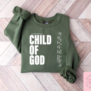 Peut inclure: Un sweat-shirt vert olive avec les mots "CHILD OF GOD" en grandes lettres blanches. Le texte "2 Corinthiens 6:18" est au-dessus du texte principal. Du texte suppl&eacute;mentaire est imprim&eacute; verticalement sur le c&ocirc;t&eacute; droit du sweat-shirt. Fond en bois clair.