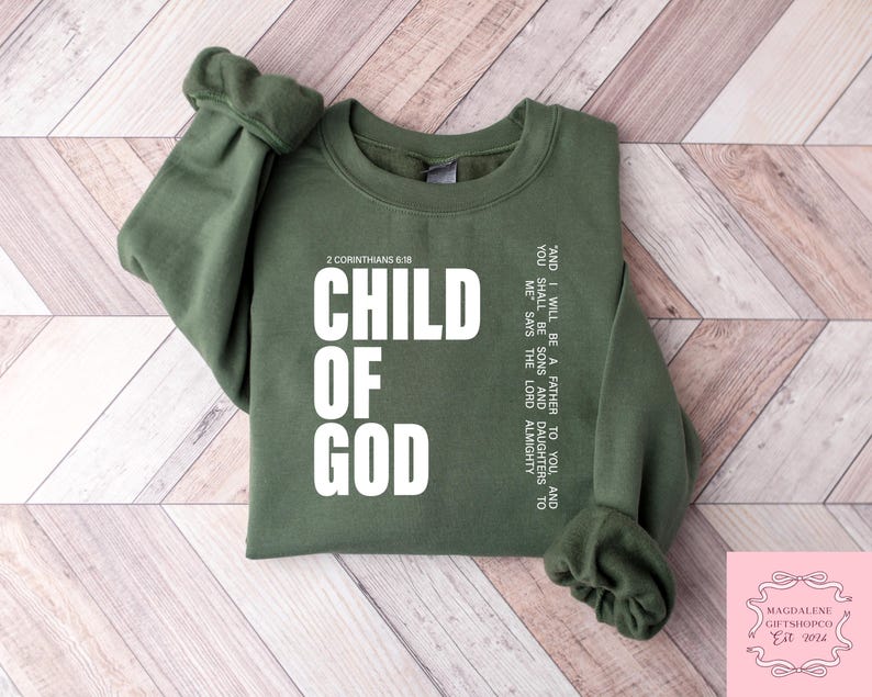 Peut inclure: Un sweat-shirt vert olive avec les mots "CHILD OF GOD" en grandes lettres blanches. Le texte "2 Corinthiens 6:18" est au-dessus du texte principal. Du texte suppl&eacute;mentaire est imprim&eacute; verticalement sur le c&ocirc;t&eacute; droit du sweat-shirt. Fond en bois clair.