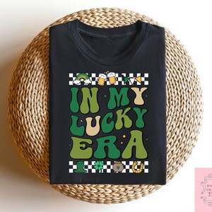 Puede incluir: Camiseta negra con un gr&aacute;fico verde, blanco y dorado que dice "In My Lucky Era". El gr&aacute;fico presenta tr&eacute;boles, jarras de cerveza y una herradura.