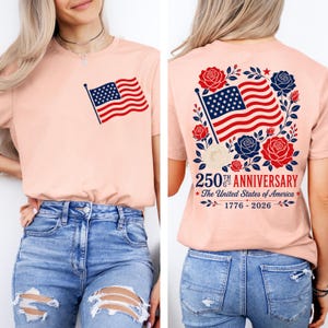 以下が含まれることがあります： 前面と背面にアメリカ国旗のデザインが施された、ピーチ色のTシャツ。背面には国旗、バラ、そして「250th Anniversary The United States of America 1776-2026」の文字が描かれています。